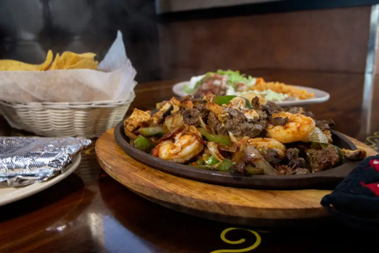 Close-up of a sizzling fajita skillet with shrimp, steak, chicken, and peppers, served with chips and side dishes on a wooden table.
