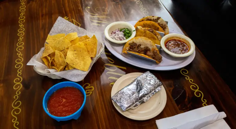Three quesabirrias served with dipping sauce, a foil-wrapped item, tortilla chips, and salsa on a wooden table.
