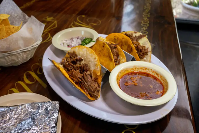 Three shredded beef tacos served with dipping sauce, diced onions, and cilantro on a platter on a wooden table.