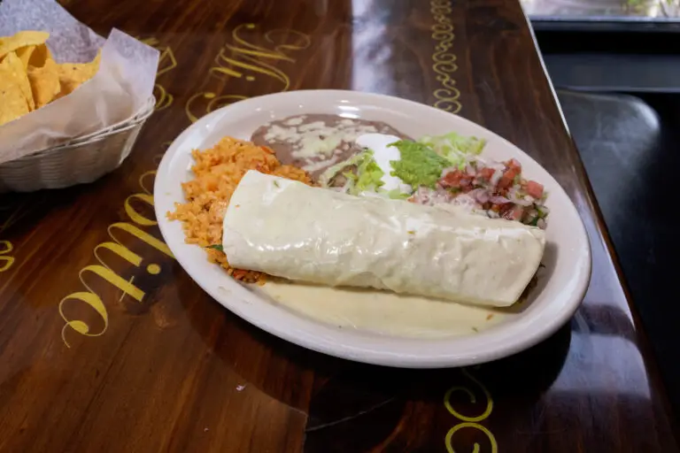 A burrito smothered in queso sauce served with Mexican rice, refried beans, guacamole, and chips on a wooden table.