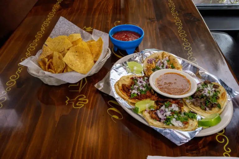 Four tacos garnished with cilantro and onions, served with lime wedges, tortilla chips, and salsa on a wooden table.