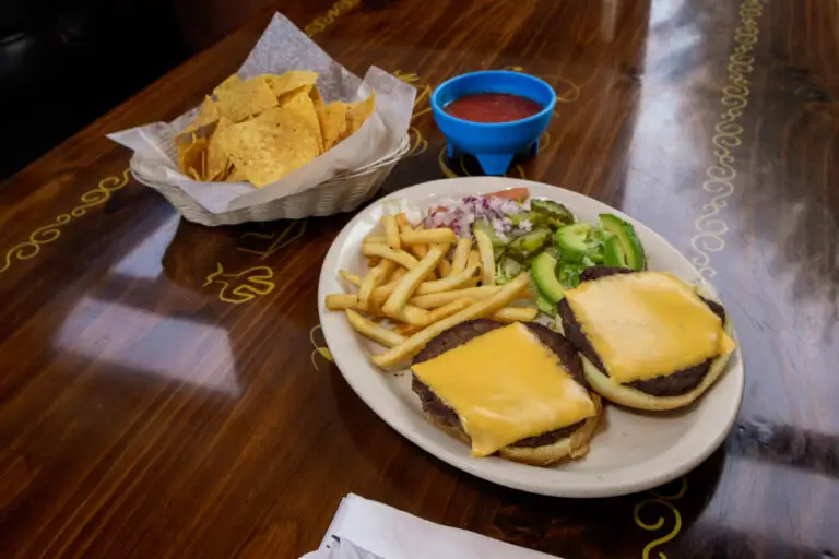 A cheeseburger served with French fries, sliced avocado, tortilla chips, and salsa on a wooden table.