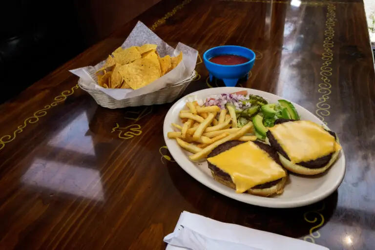 An open-faced cheeseburger served with French fries, sliced avocado, tortilla chips, and salsa on a wooden table.